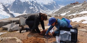 2 scientists burying a seismometer in the ground in a mountain setting