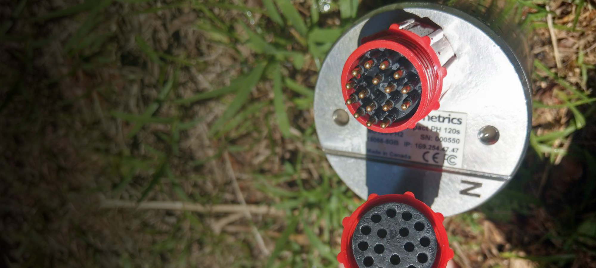 Meridian Compact posthole seismometer sitting on the grass with a red cable plug about to be connected